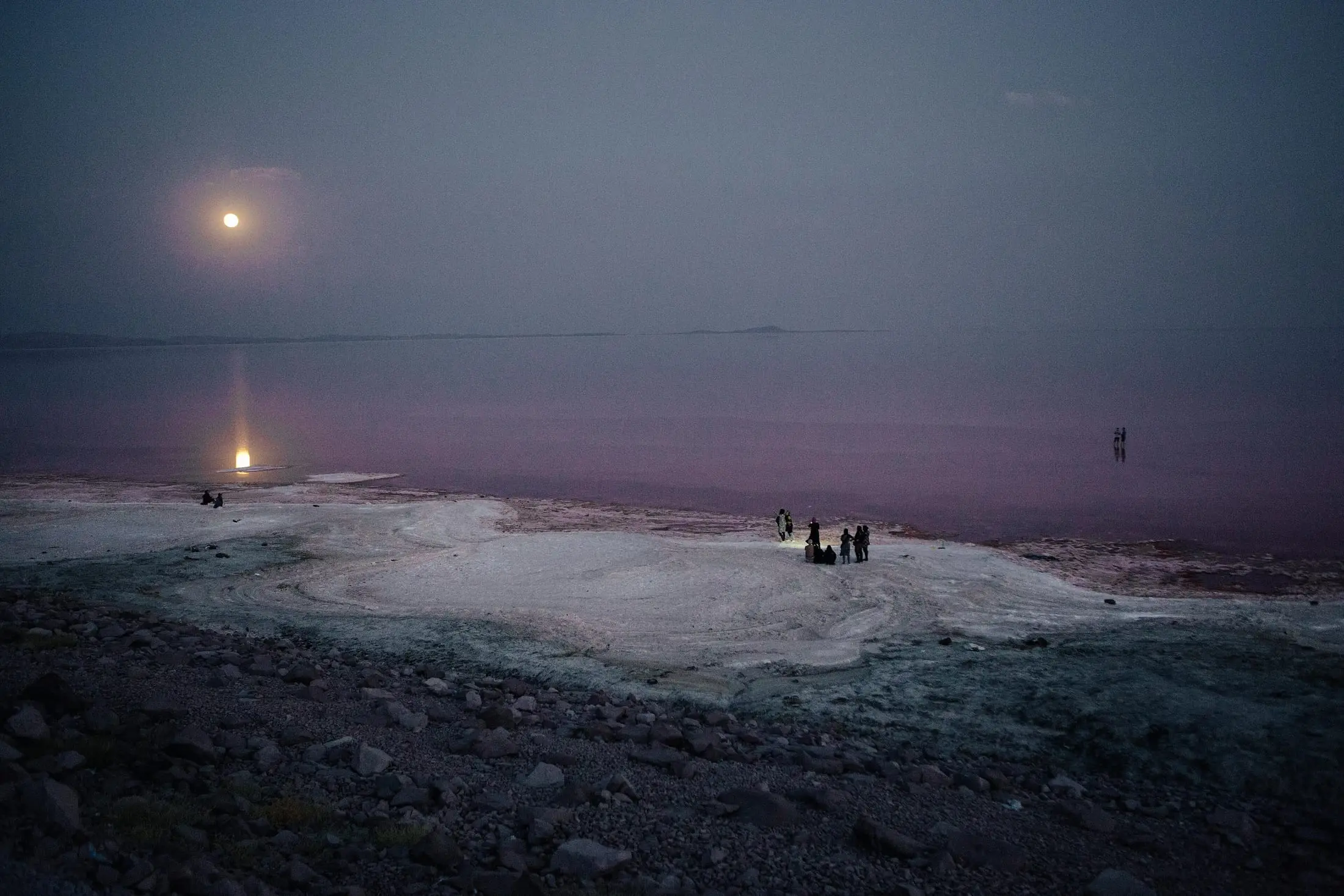 The moon rises over the salt flats of Lake Urmia at dusk; small figures stand at the water’s edge.